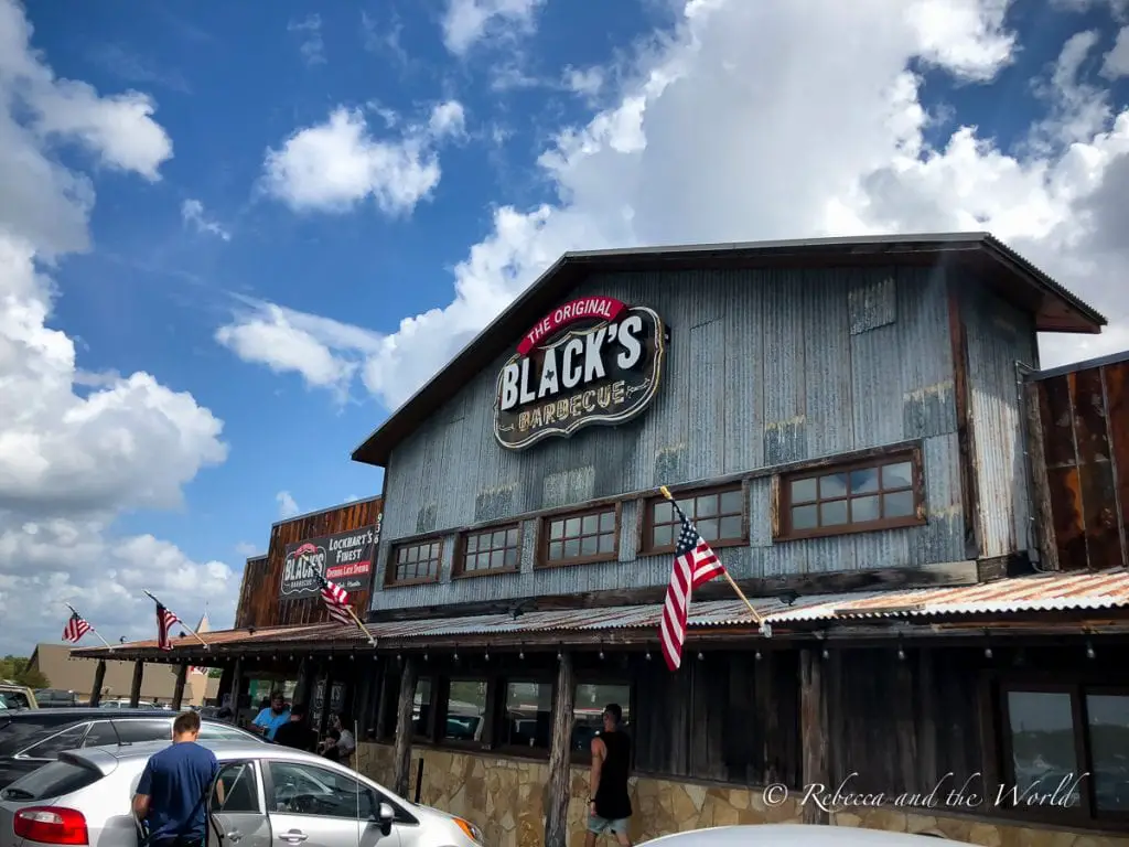 The exterior of Black's Barbecue under a cloudy sky, with rustic metal siding, American flags, and the establishment's sign visible. Black's Barbecue is a great place for lunch in New Braunfels.