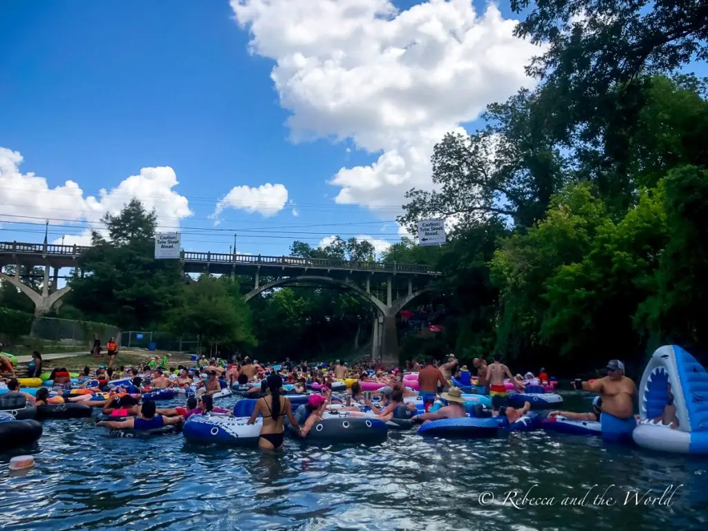 River tubing participants clustered in the water near a bridge with onlookers standing on the riverbank, under a partly cloudy sky. Tubing in Texas - in New Braunfels - is a fun way to spend a few hours.