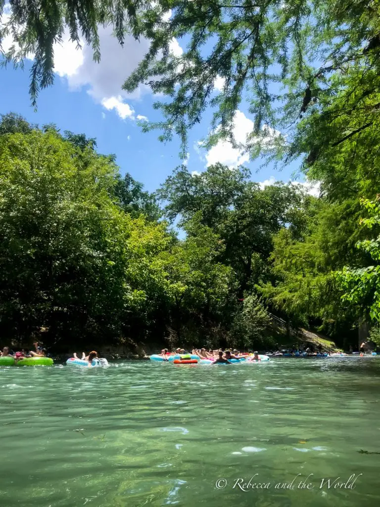 Tranquil river surrounded by dense green trees under a partly cloudy sky, with people on tubes enjoying the calm waters. The river water is spring-fed, which makes tubing in New Braunfels a refreshing thing to do on a hot summer day.