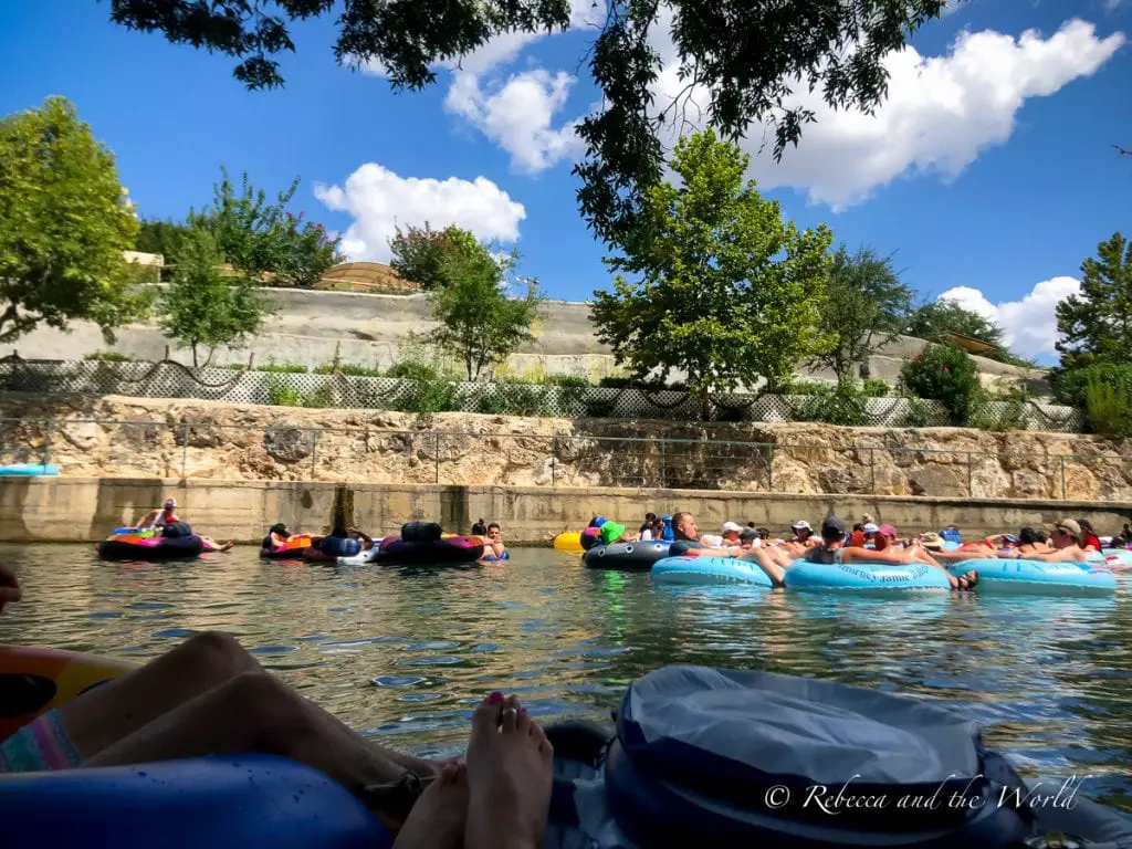 People leisurely tubing on a river with a stone wall and lush greenery on the right bank, under a clear blue sky. There are two rivers in New Braunfels for tubing - we chose the Comal River.