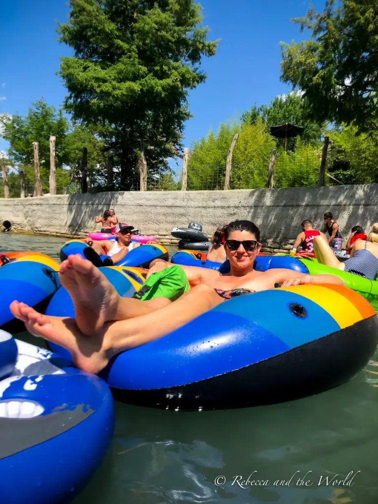 Close-up of a woman - the author of this article - reclining on a blue and yellow inner tube on the river, with other tubers and trees in the background. I had so much fun tubing in New Braunfels!