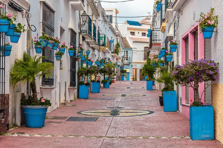 A picturesque, flower-lined street in Estepona, with vibrant blue planters and traditional Andalusian white houses adorned with colorful doors and window frames.