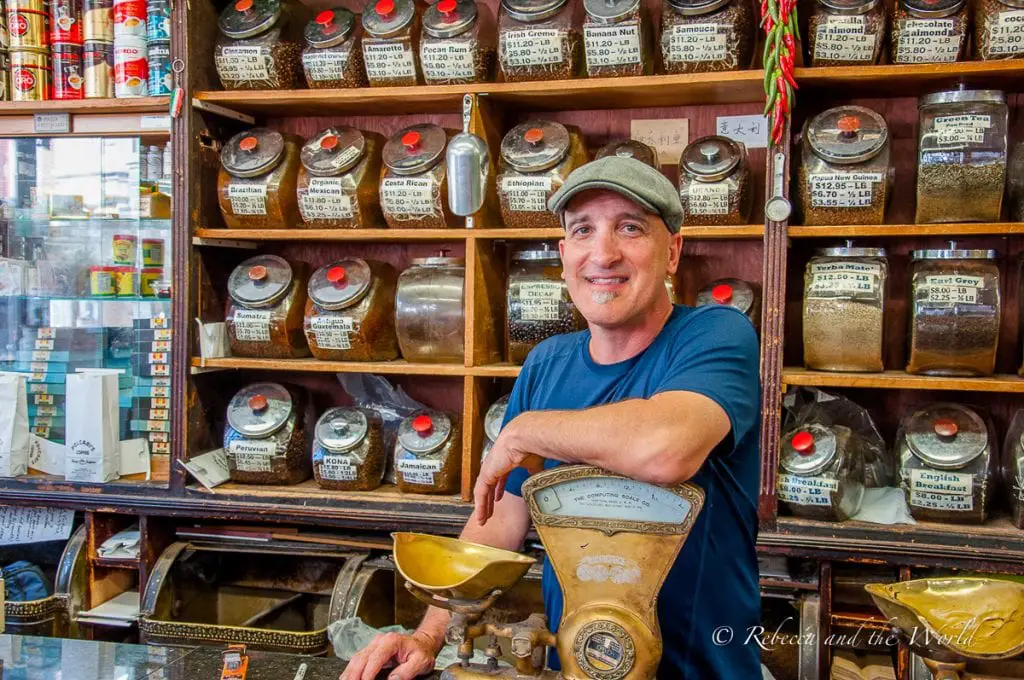 A man in a blue shirt stands smiling behind a vintage scale, surrounded by shelves filled with labeled jars of coffee and tea in a quaint shop. Bobby from Polcari's explains the history of this historic store in Boston's North End.