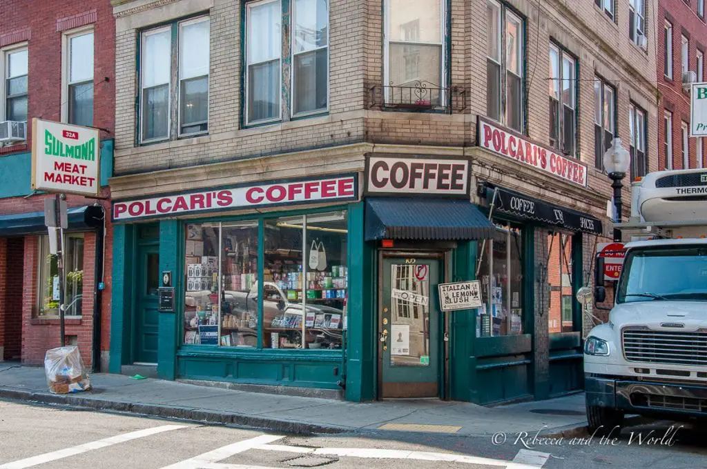 A street corner in Boston showcasing Polcari's Coffee, a classic coffee shop with a prominent red and white sign. Adjacent to it is Sulmona Meat Market with a green sign. The storefront windows display a variety of goods, suggesting a local neighborhood market atmosphere. A parked truck is visible on the right side of the frame. Polcari's is a historic store in Boston's North End.