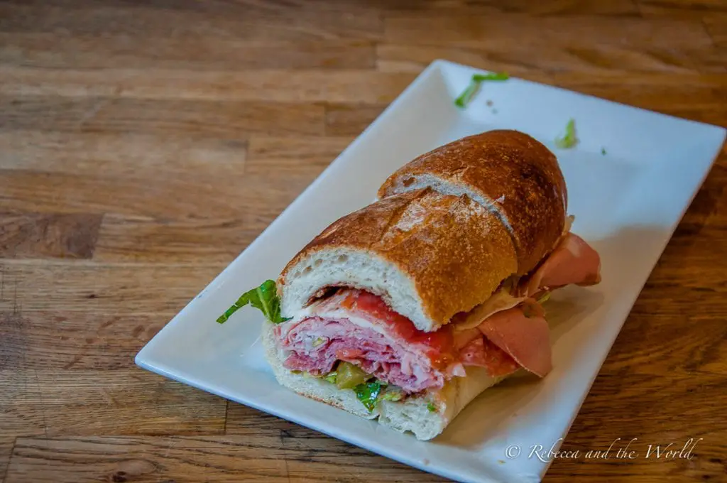 A sandwich on a white plate, cut diagonally, with Italian cold cuts, lettuce, and tomatoes on a crusty bread roll, on a wooden table. Monica's salumeria in Boston's North End is home to one of the best sandwiches I've ever eaten. Have a taste of it on this unique Boston food tour.