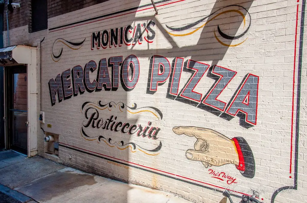 A vibrant mural on a brick wall for Monica's Mercato Pizza, featuring elaborate lettering and a pointing hand, illustrating an old-world charm. Monica's salumeria in Boston's North End is home to one of the best sandwiches I've ever eaten.