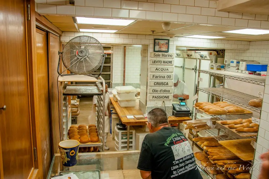 A bakery's working area with a baker in a white apron behind a counter, shelves with bread loaves, and a sign listing ingredients like "olio d'oliva" and "passione". A man in a dark-coloured t-shirt stands at the counter pointing at some of the bakery's products. The bread at Brico Panetteria is delicious.