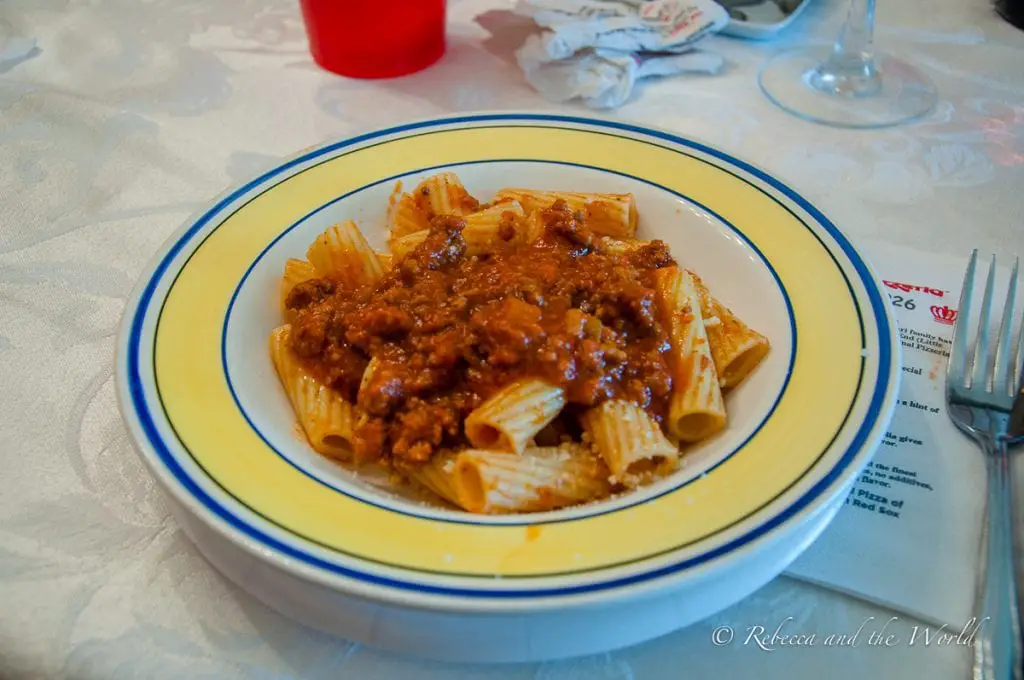 A bowl of penne pasta topped with meat sauce on a white and blue-rimmed plate, accompanied by cutlery and a red cup, suggesting a homely meal. On this Boston food tour you'll get something you can't get anywhere else - a bowl of Mama Maria's rigatoni with Bolognese sauce.