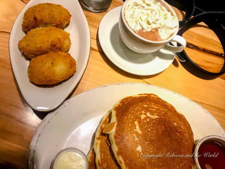 A breakfast spread with golden-brown pancakes, whipped hot chocolate, and crispy fried potato cakes on white ceramic dishes.