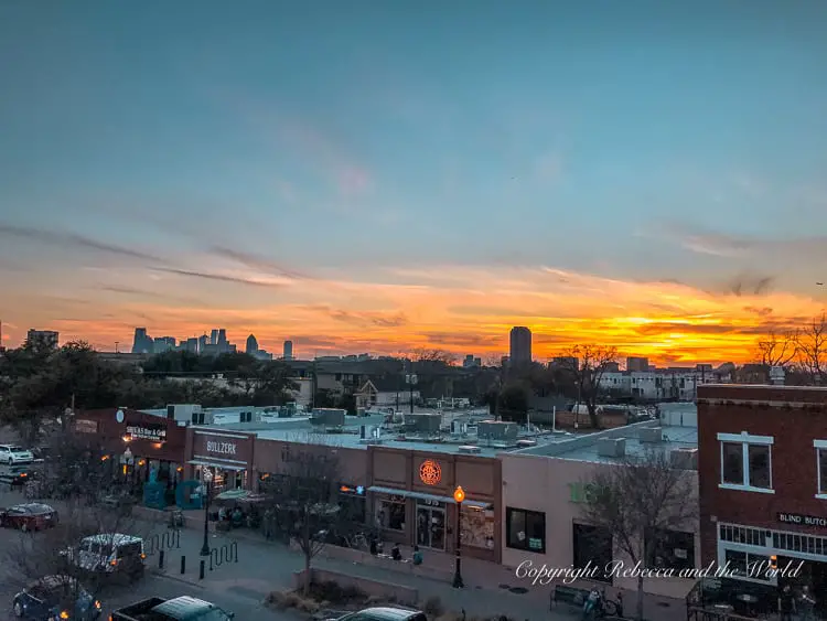 A sunset view over the rooftops of the Lower Greenville neighborhood in Dallas, with the city skyline silhouetted against a vibrant sky.