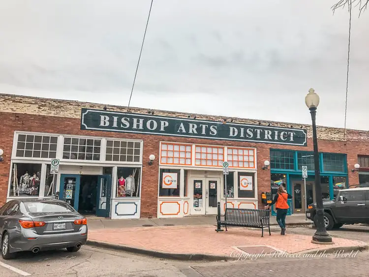 The front of shops and eateries with 'Bishop Arts District' signage, under an overcast sky with pedestrians and parked cars.