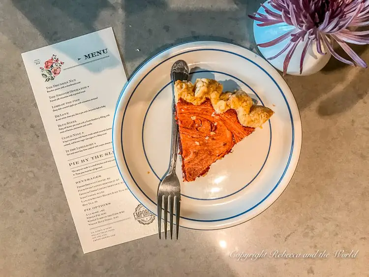 A slice of pie on a white plate with a blue rim, a fork, and a menu on a marbled table. You must try Emporium Pies on your weekend in Dallas.