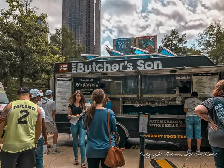 People queued at 'The Butcher's Son' food truck, ordering gourmet food on a sunny day with trees and a high-rise building nearby. Klyde Warren Park is one of the best things to do in Dallas, and a must for your itinerary.