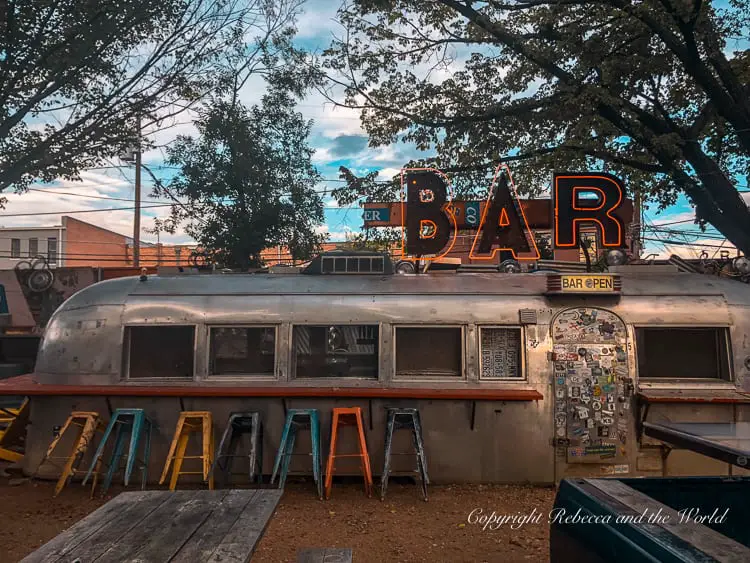 An outdoor bar setting with colorful stools in front of a vintage airstream trailer, under the glow of a neon 'BAR' sign at dusk.