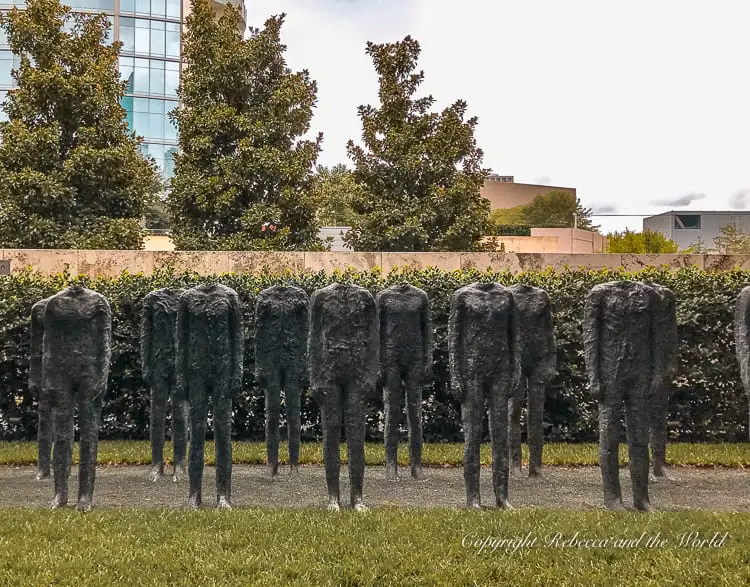 Sculpture installation of numerous dark figures with no heads, standing in rows on a lawn, with greenery and a high-rise building in the background. The Nasher Sculpture Center is one of the best museums in Dallas.