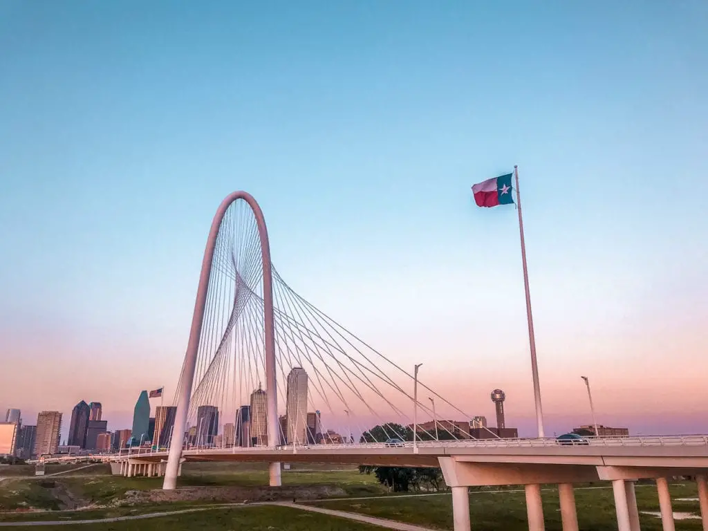 The Margaret Hunt Hill Bridge in Dallas with its distinctive arch spanning the Trinity River at sunset, with the Dallas skyline in the background and the Texas flag in the foreground.