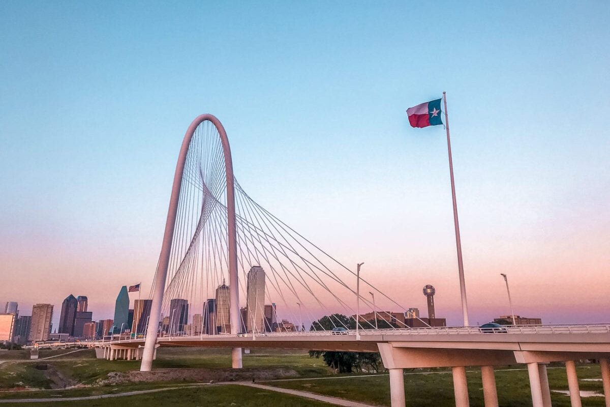 The Margaret Hunt Hill Bridge in Dallas with its distinctive arch spanning the Trinity River at sunset, with the Dallas skyline in the background and the Texas flag in the foreground.