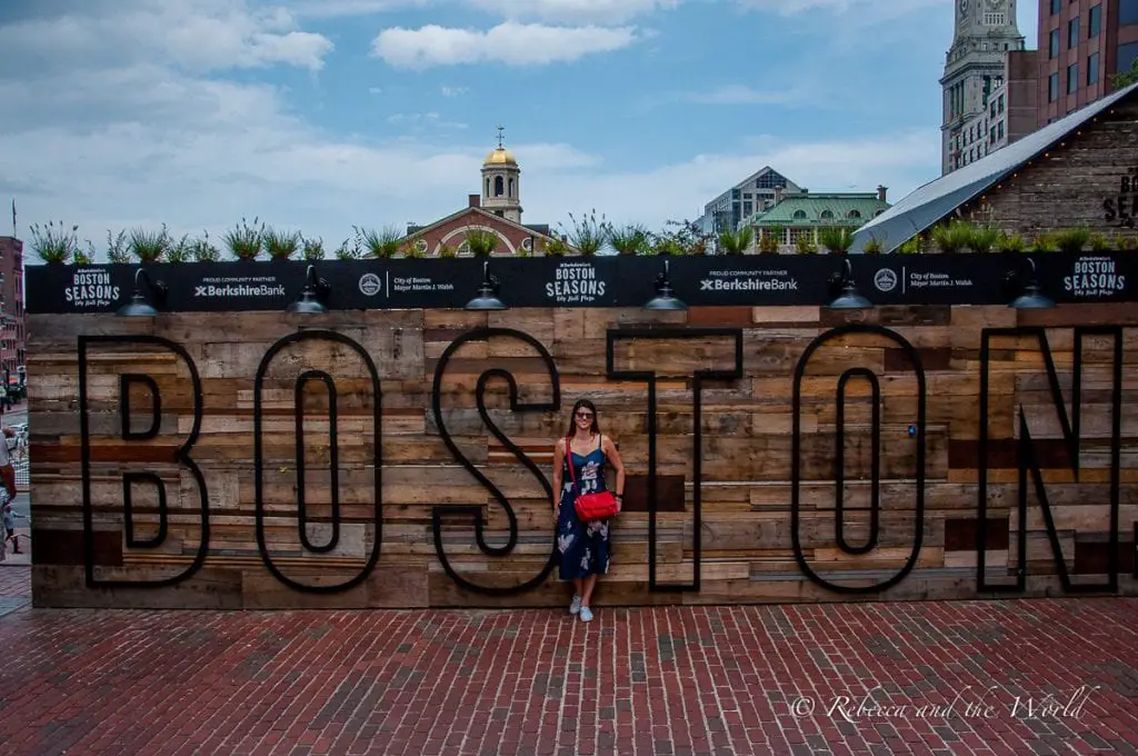 A woman - the author of this article - standing in front of a large wooden sign with the word "BOSTON" in bold letters, backed by city buildings and clear skies. Boston food is a great reason to visit the city!