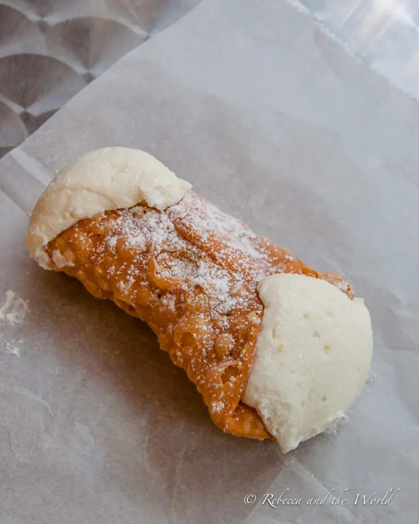 A close-up of a cannoli with creamy filling spilling out from the crispy shell, sitting on a piece of parchment paper. There's a "cannoli war" between Mike's Pasty and Modern Pastry in Boston - try both to see which one you prefer!