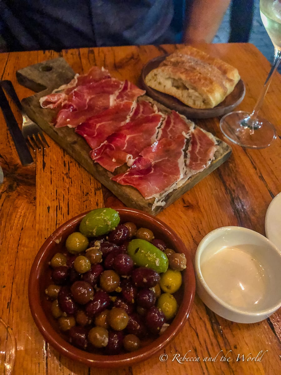 A rustic wooden board with slices of cured meats, a side of bread, a bowl of mixed olives, and a small dish of sauce, with a glass of white wine in the background. The tapas at Toro in Boston are delicious.