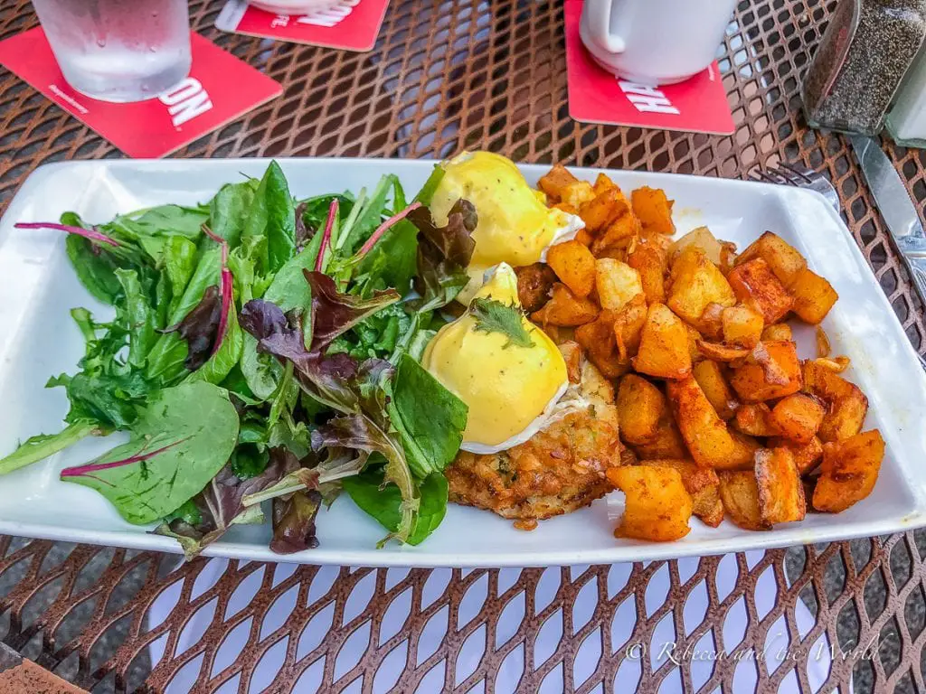 A brunch plate featuring eggs Benedict with hollandaise sauce, seasoned home fries, and a side of fresh green salad, on an outdoor table with coasters and a frosted glass. The crab cake Benedict from Stephanie's is a great brunch choice.