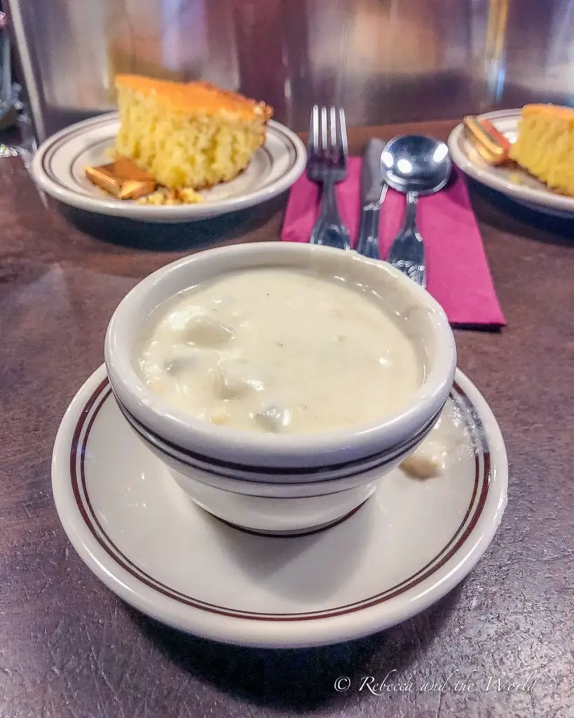 A bowl of creamy clam chowder served alongside a slice of cornbread, presented on a restaurant table. I was surprised that I liked clam chowder so much - it's delicious!