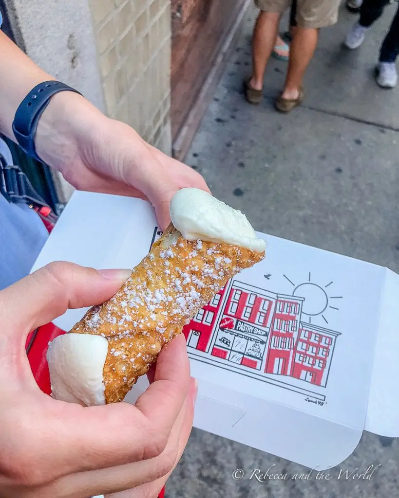 A person's hand holding a cannoli with two scoops of white filling at each end, dusted with powdered sugar, with a pastry shop illustrated on the box. Cannoli is one of the best foods in Boston that you have to try when you visit.