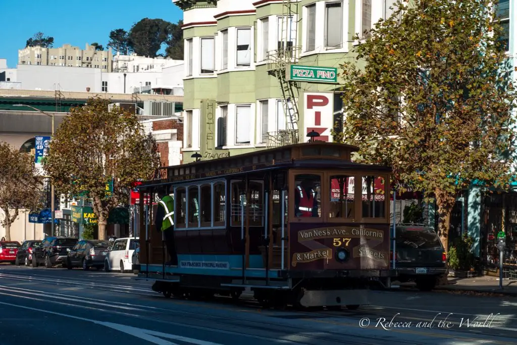 A traditional San Francisco cable car marked "Van Ness Ave. California St. & Market St." on a city street with buildings and trees in the background, and a cable car operator standing on the running board. San Francisco's cable cars are a fun and easy way to get up and down the city's steep hills.