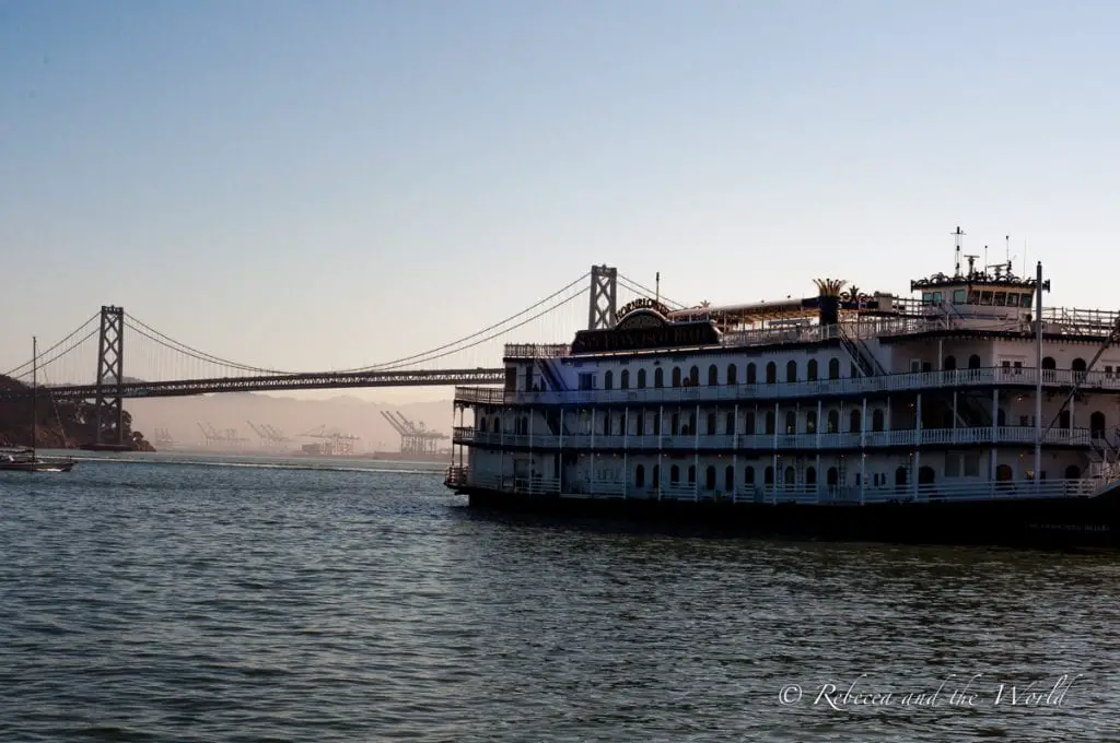 A white, multi-decked riverboat named 'San Francisco Belle' docked on calm waters, with the Golden Gate Bridge in the background partially obscured by mist and the setting sun. Every San Francisco itinerary should include a wander along the Embarcadero.