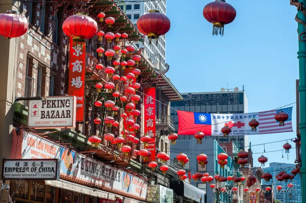 A busy street in Chinatown with red lanterns strung across the road, colorful storefront banners, and a mix of traditional and modern buildings under a clear sky. You must include a visit to Chinatown on your 2 days in San Francisco itinerary.