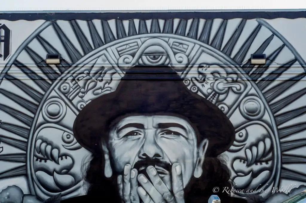 A detailed black and white mural of a person's face with expressive eyes and a hand to the lips, set against an intricate Aztec-inspired pattern. The Santana mural in the Mission District in San Francisco is so lifelike.