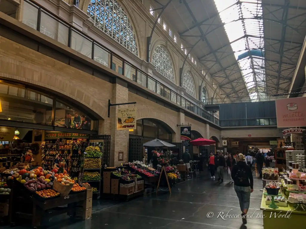 Indoor view of a bustling market hall with high arched ceilings and natural light streaming through. Various stalls display fresh produce and baked goods, with people browsing the offerings. The Ferry Building in San Francisco is a great place to start your itinerary.