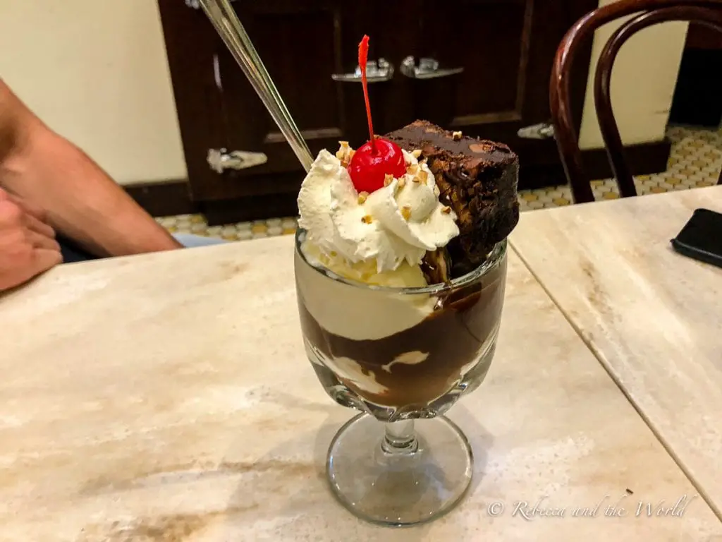 Close-up of a sundae glass filled with vanilla ice cream, chocolate sauce, whipped cream, a cherry on top, and a large brownie piece. The glass sits on a marble table with a person partially visible in the background. One of the best things to eat in San Francisco is a delectable ice cream sundae from Ghirardelli.