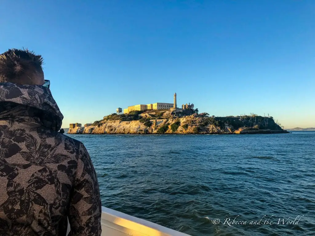 View from a boat of Alcatraz Island at sunset, with a figure in a patterned jacket observing the island. The prison complex is illuminated by the warm light of the setting sun. Only got 2 days in San Francisco? Then make sure a visit to Alcatraz is included.