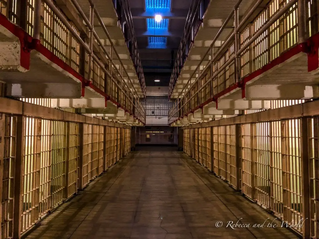 Long, eerie corridor inside Alcatraz prison showing two levels of cells with barred doors, dimly lit by overhead lights and natural light filtering through the skylights. Only got 2 days in San Francisco? Then make sure a visit to Alcatraz is included. An Alcatraz night tour is the best option!