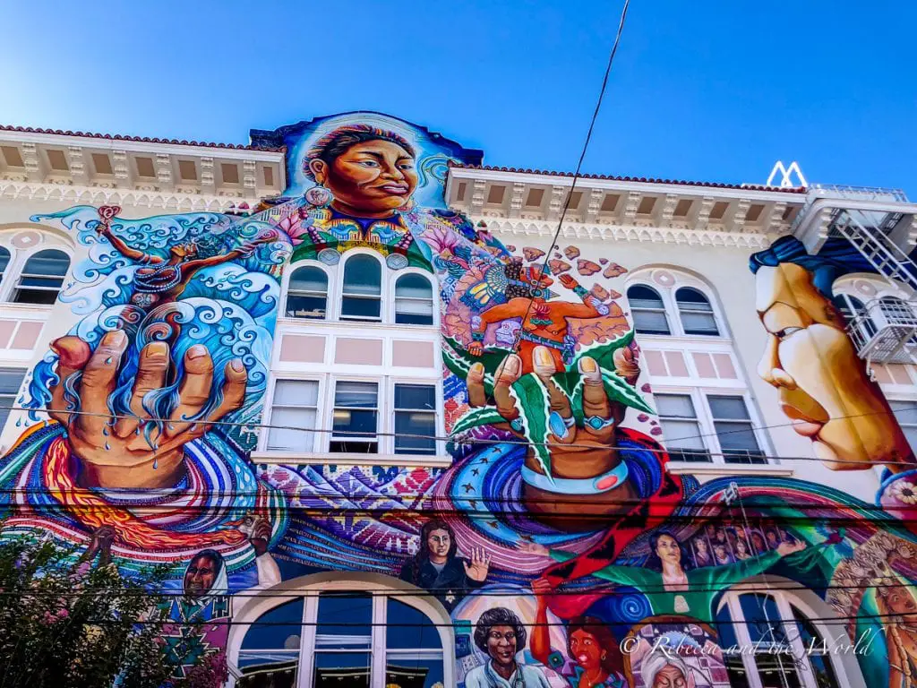 A vibrant street mural depicting a large, colourful female figure with extended hands and a background filled with faces and cultural symbols. One of the most beautiful murals in San Francisco is this one on the side of the Women's Building in the Mission District.