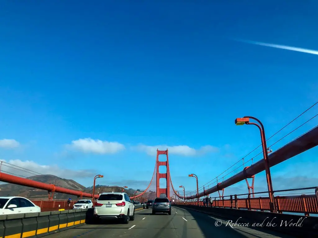 Driving perspective on the Golden Gate Bridge with traffic and the distinctive red railing, heading towards the Marin Headlands under a bright blue sky. Heading to Sonoma from San Francisco includes a drive across the Golden Gate Bridge.