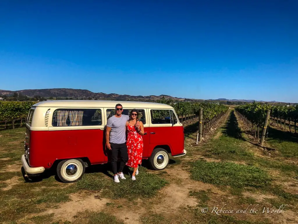 A woman in red (the author) and a man in grey t-shirt stand in front of a red and white Combi in front of vineyards in Sonoma, California