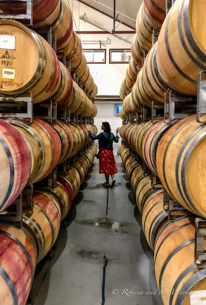 Inside a winery's barrel room, a woman - the author of this article - in a red dress and denim jacket is seen from behind, walking between towering stacks of wooden wine barrels arranged in parallel lines. Winery tours in Sonoma allow a behind the scenes look at some of the best wineries in Sonoma.