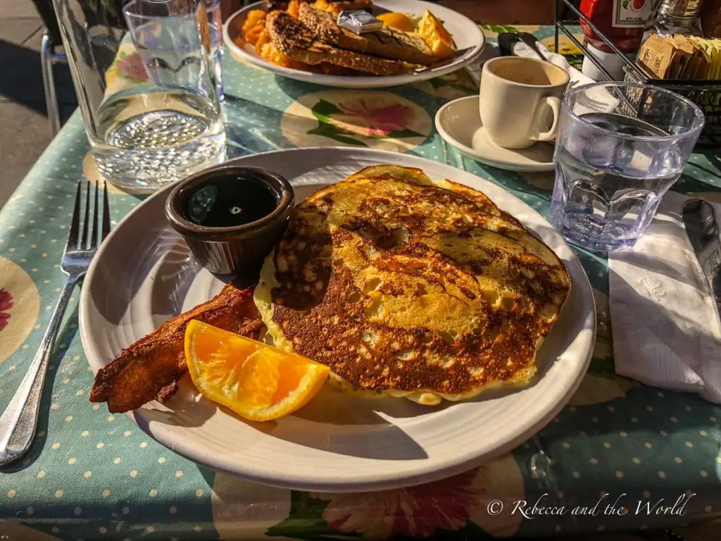 A breakfast plate with a large pancake, a side of bacon, an orange slice, and a small cup of syrup, accompanied by a coffee cup and a water glass, on a table with a polka-dotted tablecloth. Try the zucchini pancakes at Mymy in San Francisco.