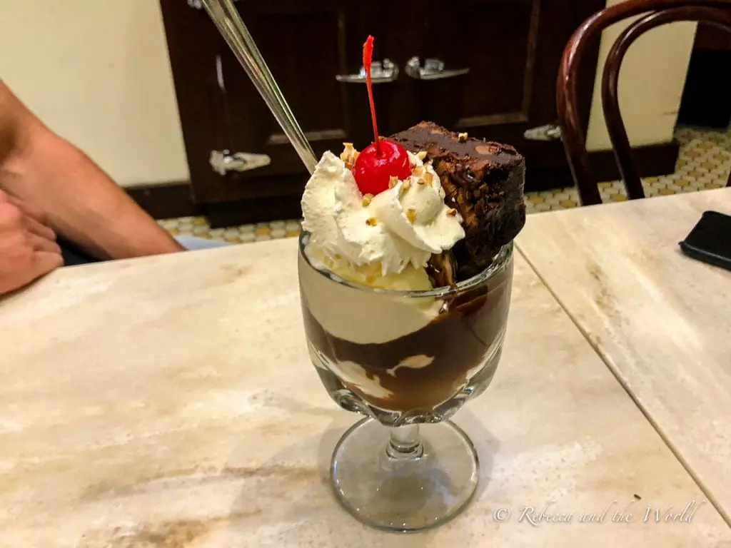 An indulgent dessert with layers of chocolate, vanilla ice cream, whipped cream, and a cherry on top, served in a tall glass with a spoon. Stop by Ghirardelli in San Francisco to try the amazing ice cream sundae!