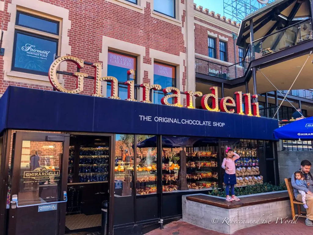 The facade of the "Ghirardelli" chocolate shop with large lit-up lettering, seen during daylight with shoppers passing by. Stop by Ghirardelli in San Francisco to try the amazing ice cream sundae!