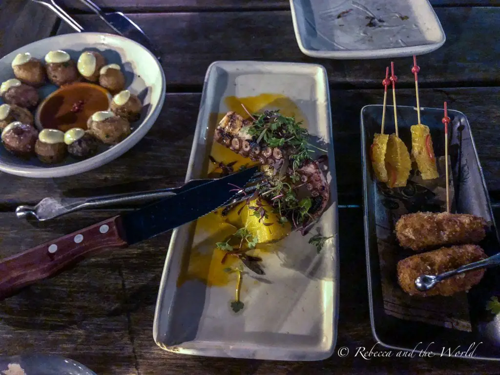 An evening dining scene featuring small plates of various foods including patatas bravas, grilled octopus, and fried croquettes on a dark wooden table. The tapas at Spanish restaurant Coqueta in San Francisco are a must.
