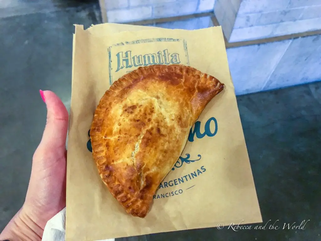A hand holding a brown paper bag with "Humita" printed on it, containing a golden-brown, crescent-shaped pastry called an empanada. The empanadas at El Porteno in San Francisco are delicious.