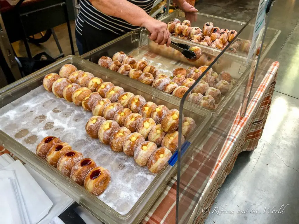 A clear display case containing rows of sugar-covered donuts, some filled with cream and others with a red fruit jam, being served by a person in a striped shirt. So many donuts to try at the Ferry Building in San Francisco!