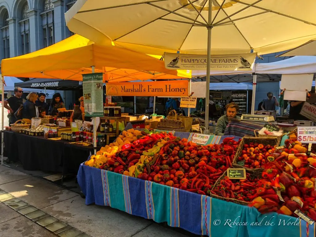 A bustling outdoor farmers' market with a vibrant display of assorted vegetables and fruits, with various vendor stalls under yellow umbrellas. The Farmers Market at the Ferry Building in San Francisco is great for local produce.