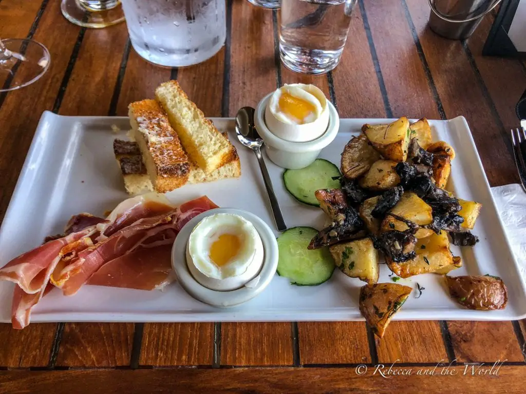 A brunch plate featuring slices of toasted bread, a soft-boiled egg in an egg cup, slices of prosciutto, roasted potatoes with mushrooms, and cucumber slices, all arranged on a rectangular white plate on a wooden table. Stop in Sausalito on your way to Sonoma, and grab brunch at Le Garage.