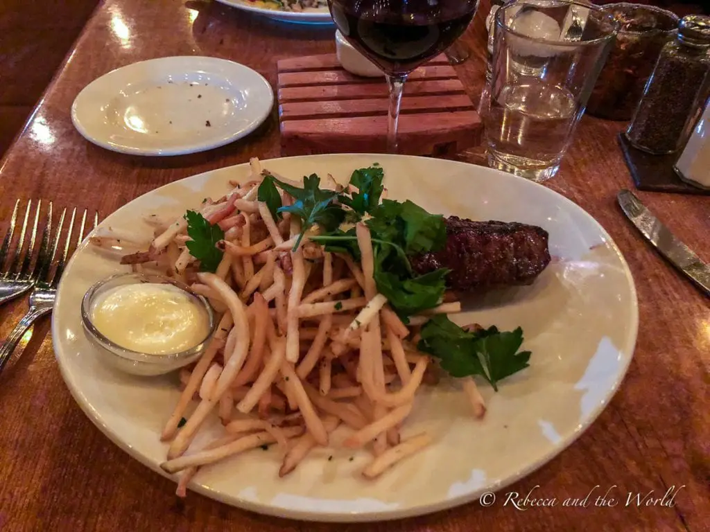 A plate of steak frites, with a grilled steak garnished with parsley beside a pile of thin, crispy French fries, served with a side of mayonnaise, on a round white plate, with a wine glass and water glass in the background. Farm-to-table restaurant the girl and the fig in Sonoma is one of the best restaurants in Sonoma.