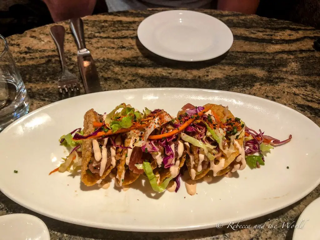 A gourmet dish of crispy wanton "tacos" filled with tuna and topped with a colourful slaw, drizzled with a creamy sauce, presented on an oval white plate on a granite countertop. Grab a casual meal at El Dorado Kitchen in Sonoma.