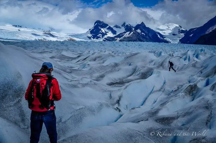 A man in a red jacket with a red backpack walks on Perito Moreno Glacier, with blue ice caps that are dirty in parts. Another man in dark clothes walks further away. Perito Moreno is a must visit on an Argentina 2 week itinerary